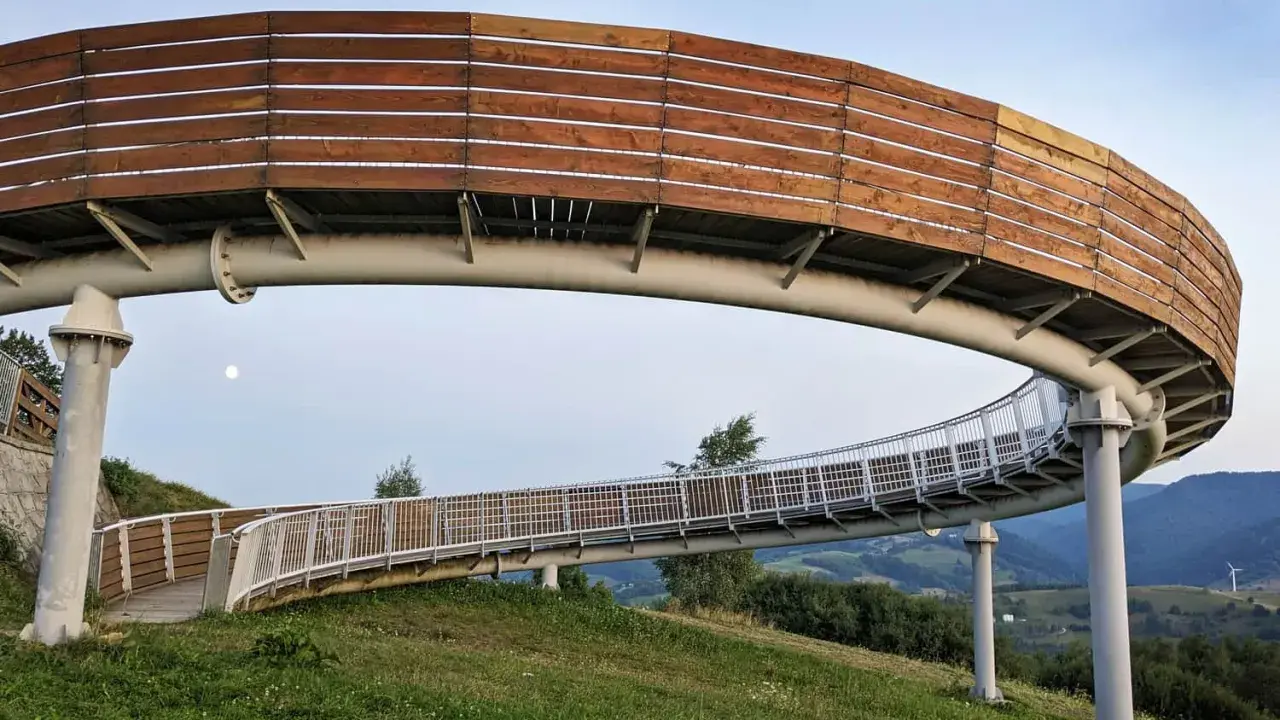 Drewniana kładka widokowa wznosi się nad zielonymi wzgórzami, oferując panoramę jak z **Woli Kroguleckiej**. Księżyc w pełni towarzyszy spacerowiczom.