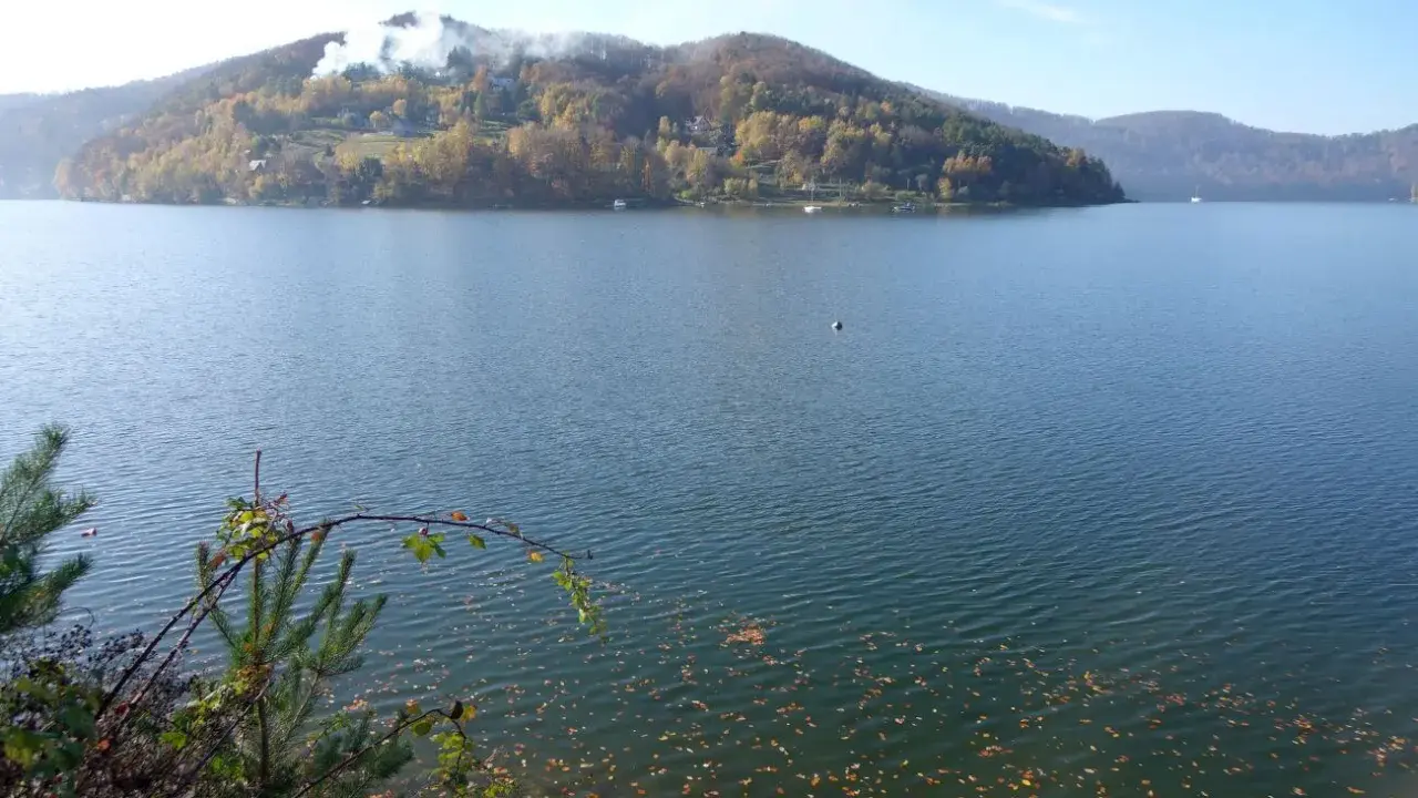 Jesioro Rożnowskie jesienią. Woda spokojna, na wzgórzach złote liście, a na szczycie widać dym z kominów.
