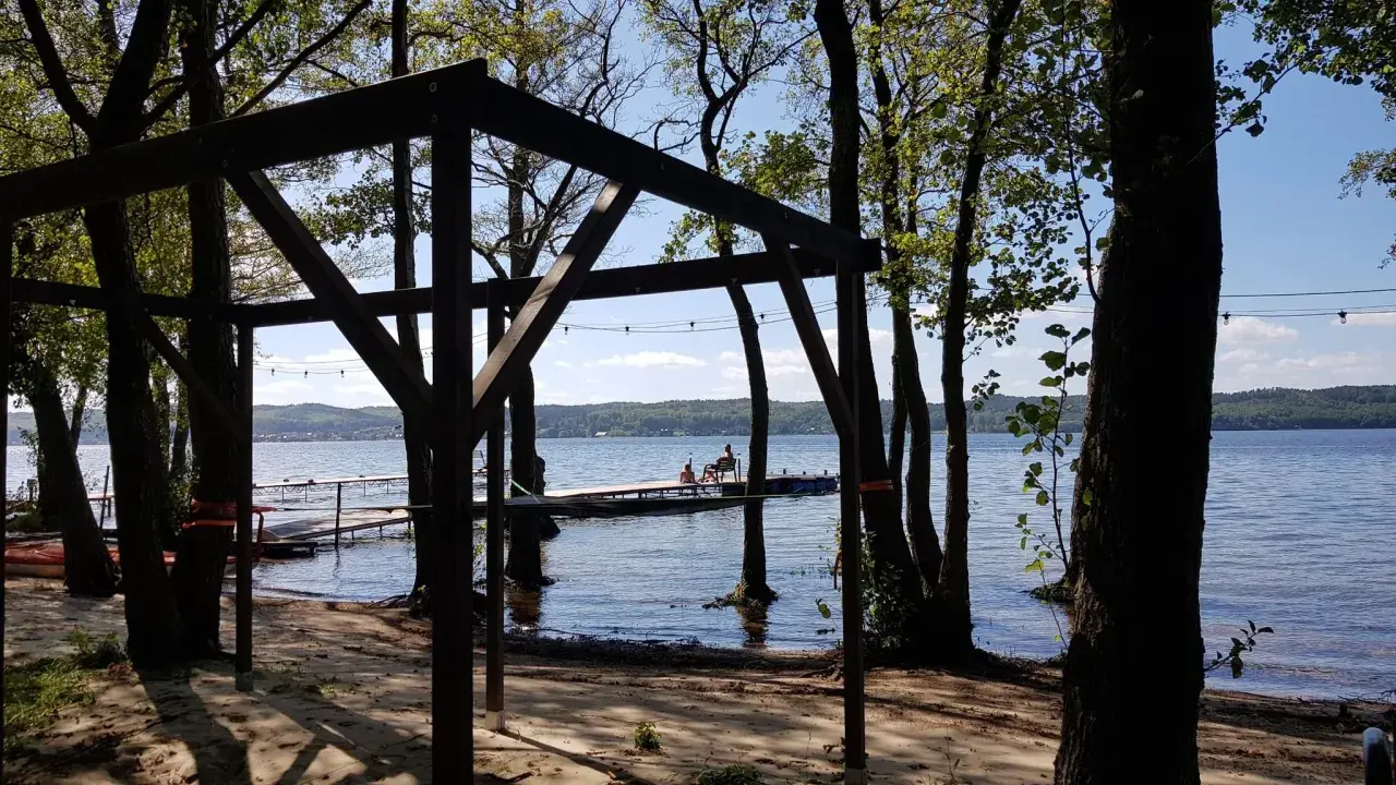 Drewniana konstrukcja na plaży, z widokiem na jezioro i pomost, gdzie siedzą ludzie. Wokół drzewa, tworzące naturalne atrakcje.