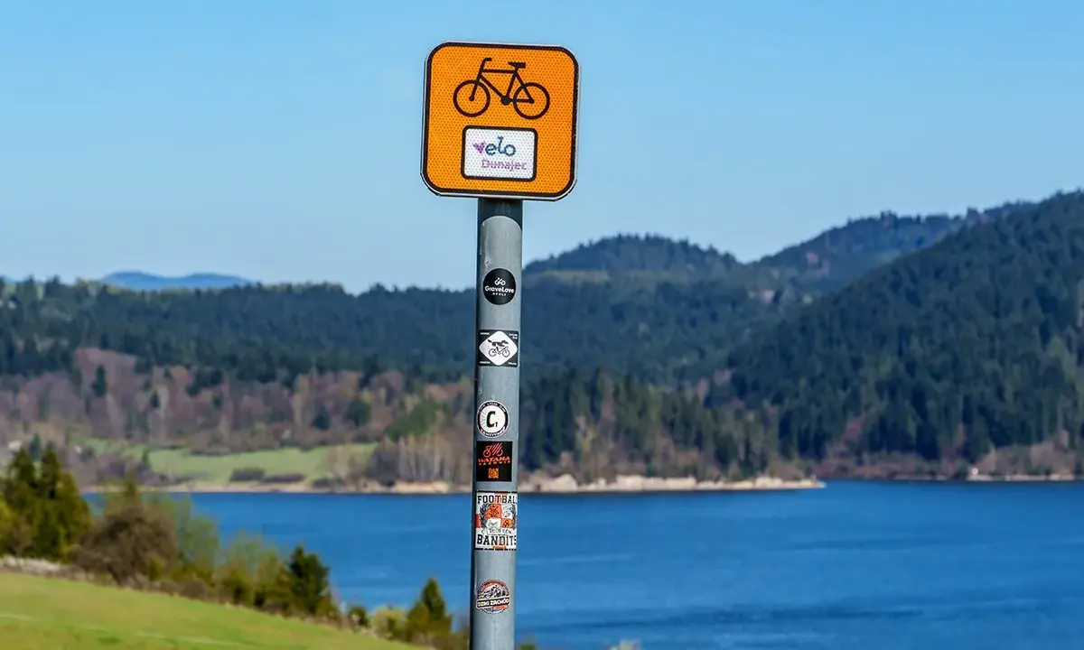 Znak Velo Dunajec zapowiada piękną trasę rowerową. Jezioro i góry tworzą malowniczy krajobraz.