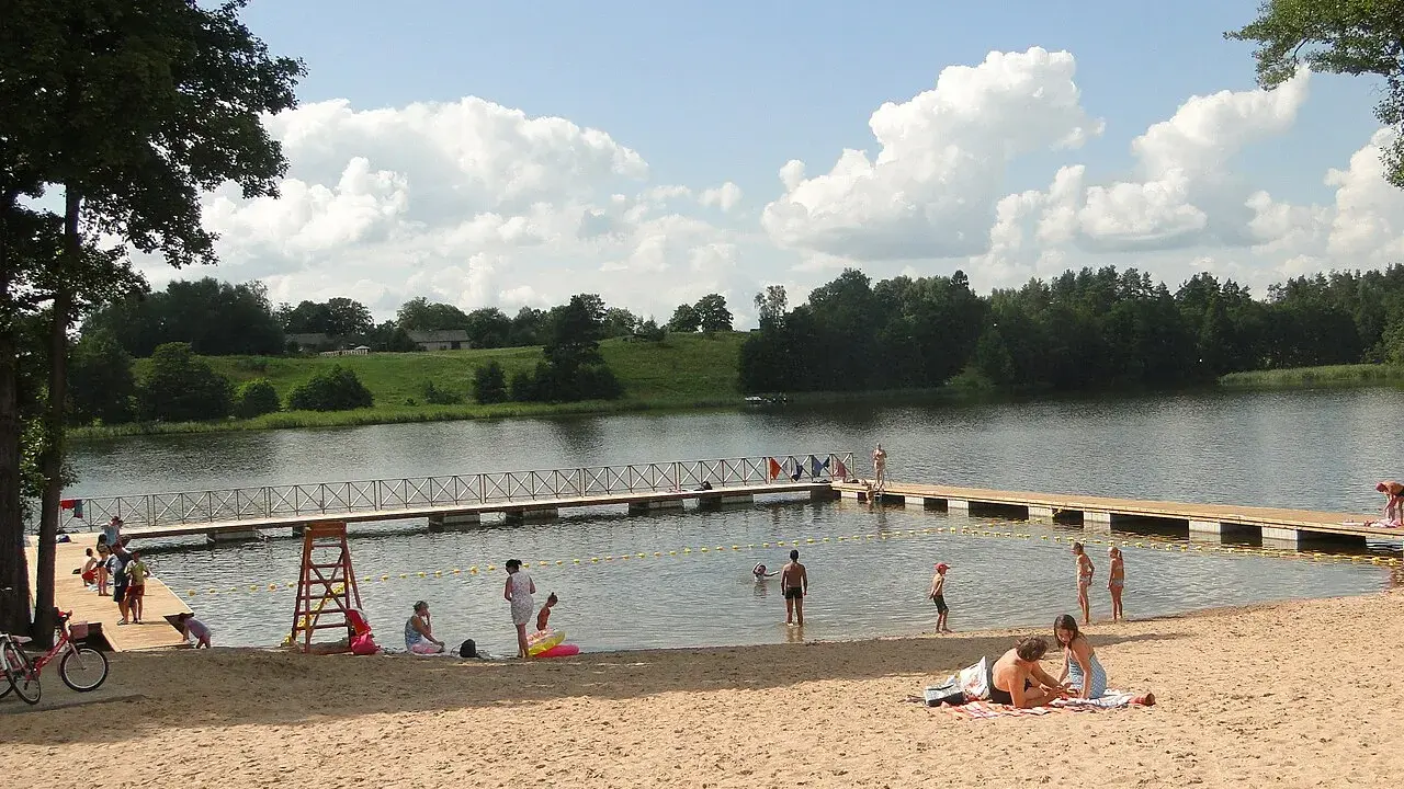 Słoneczna mazury plaża z ludźmi odpoczywającymi na piasku i kąpiącymi się w jeziorze. Drewniane molo w wodzie.