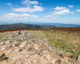 Kamienista trasa na Śnieżniku z widokiem na góry i chmury. Dwie osoby wędrują po szlaku.