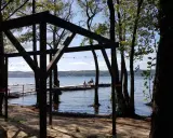 Drewniana konstrukcja na plaży, z widokiem na jezioro i pomost, gdzie siedzą ludzie. Wokół drzewa, tworzące naturalne atrakcje.