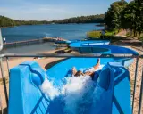 Chłopiec zjeżdża po niebieskiej zjeżdżalni wodnej, wpadając do jeziora Dziemiany. W tle widać plażę i las.