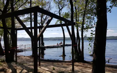 Drewniana konstrukcja na plaży, z widokiem na jezioro i pomost, gdzie siedzą ludzie. Wokół drzewa, tworzące naturalne atrakcje.