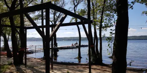 Drewniana konstrukcja na plaży, z widokiem na jezioro i pomost, gdzie siedzą ludzie. Wokół drzewa, tworzące naturalne atrakcje.