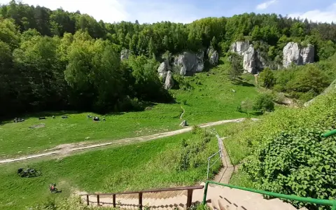 Zielona dolina kobylańska z wapiennymi skałami, lasem i ludźmi odpoczywającymi na trawie. Schody prowadzą w dół.