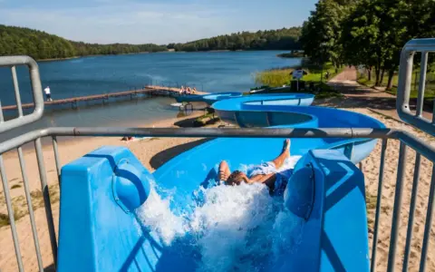 Chłopiec zjeżdża po niebieskiej zjeżdżalni wodnej, wpadając do jeziora Dziemiany. W tle widać plażę i las.