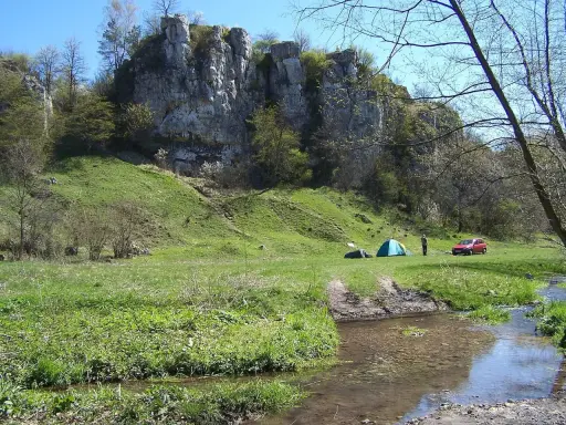 Malownicza dolina Kluczwody z rozbitymi namiotami, czerwonym samochodem i strumieniem. Skaliste zbocza porośnięte zielenią.