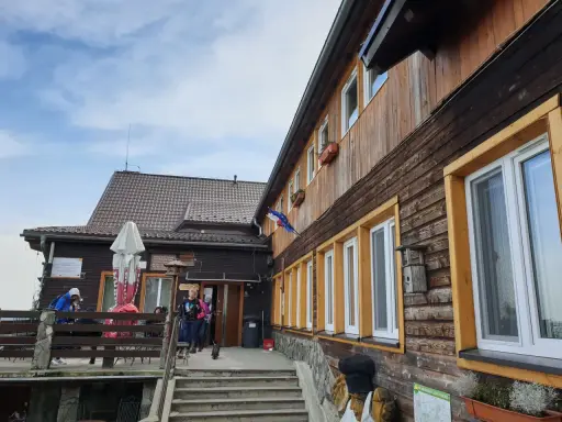 Drewniane schronisko w Górach Opawskich, z tarasem i schodami. W tle błękitne niebo z chmurami.