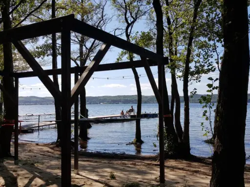 Drewniana konstrukcja na plaży, z widokiem na jezioro i pomost, gdzie siedzą ludzie. Wokół drzewa, tworzące naturalne atrakcje.