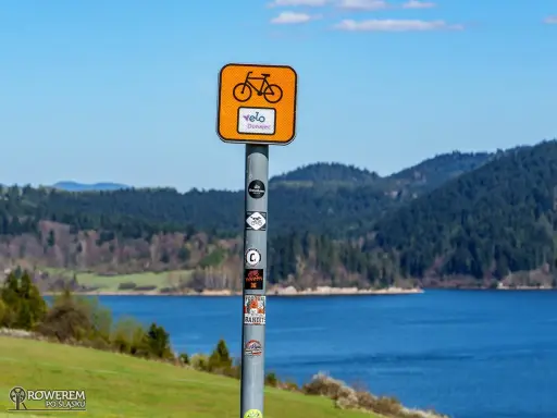 Znak Velo Dunajec zapowiada piękną trasę rowerową. Jezioro i góry tworzą malowniczy krajobraz.