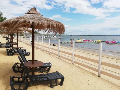 Plaża nad Jeziorem Nyskim z leżakami i parasolami. W tle widać wodę, pomost z łódkami i statek wycieczkowy. Idealne miejsce na wypoczynek i odkrywanie atrakcji.