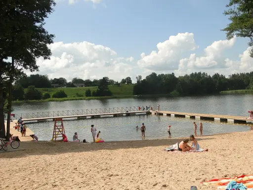 Słoneczna mazury plaża z ludźmi odpoczywającymi na piasku i kąpiącymi się w jeziorze. Drewniane molo w wodzie.