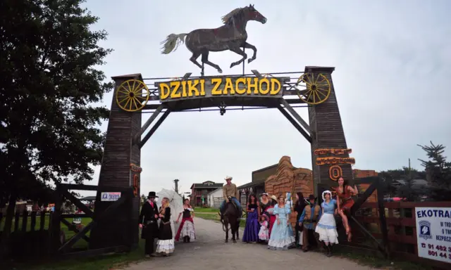 Park rozrywki Dziki Zachód koło Kołobrzegu. Grupa osób w strojach z Dzikiego Zachodu pozuje przed bramą.