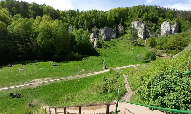 Zielona dolina kobylańska z wapiennymi skałami, lasem i ludźmi odpoczywającymi na trawie. Schody prowadzą w dół.