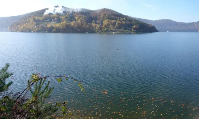 Jesioro Rożnowskie jesienią. Woda spokojna, na wzgórzach złote liście, a na szczycie widać dym z kominów.