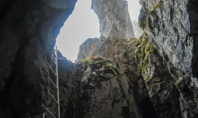 Wąska szczelina skalna, przez którą widać niebo. Metalowa drabina prowadzi w górę. To wejście do jaskini raptawickiej.