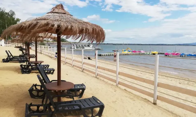 Plaża nad Jeziorem Nyskim z leżakami i parasolami. W tle widać wodę, pomost z łódkami i statek wycieczkowy. Idealne miejsce na wypoczynek i odkrywanie atrakcji.