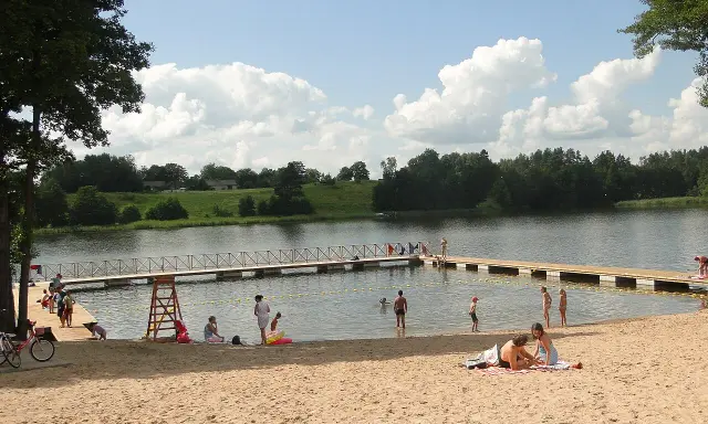 Słoneczna mazury plaża z ludźmi odpoczywającymi na piasku i kąpiącymi się w jeziorze. Drewniane molo w wodzie.