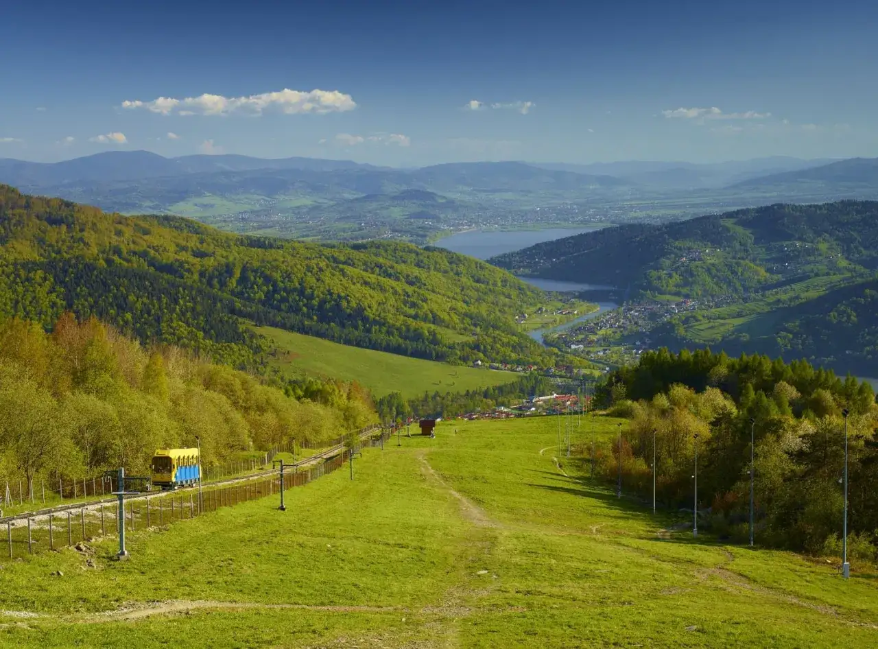 Widok na Beskid Mały z kolejką górską. Zielone wzgórza, jezioro i miasteczko w dolinie.